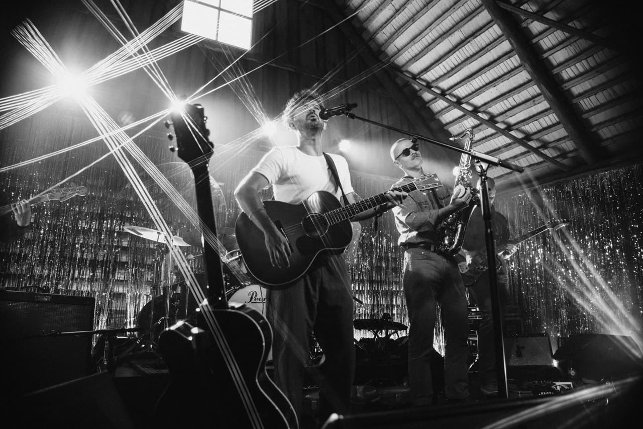 Araukaaria live on stage: Pepi singing with an acoustic guitar, a saxophonist beside him, under a wooden barn roof.
