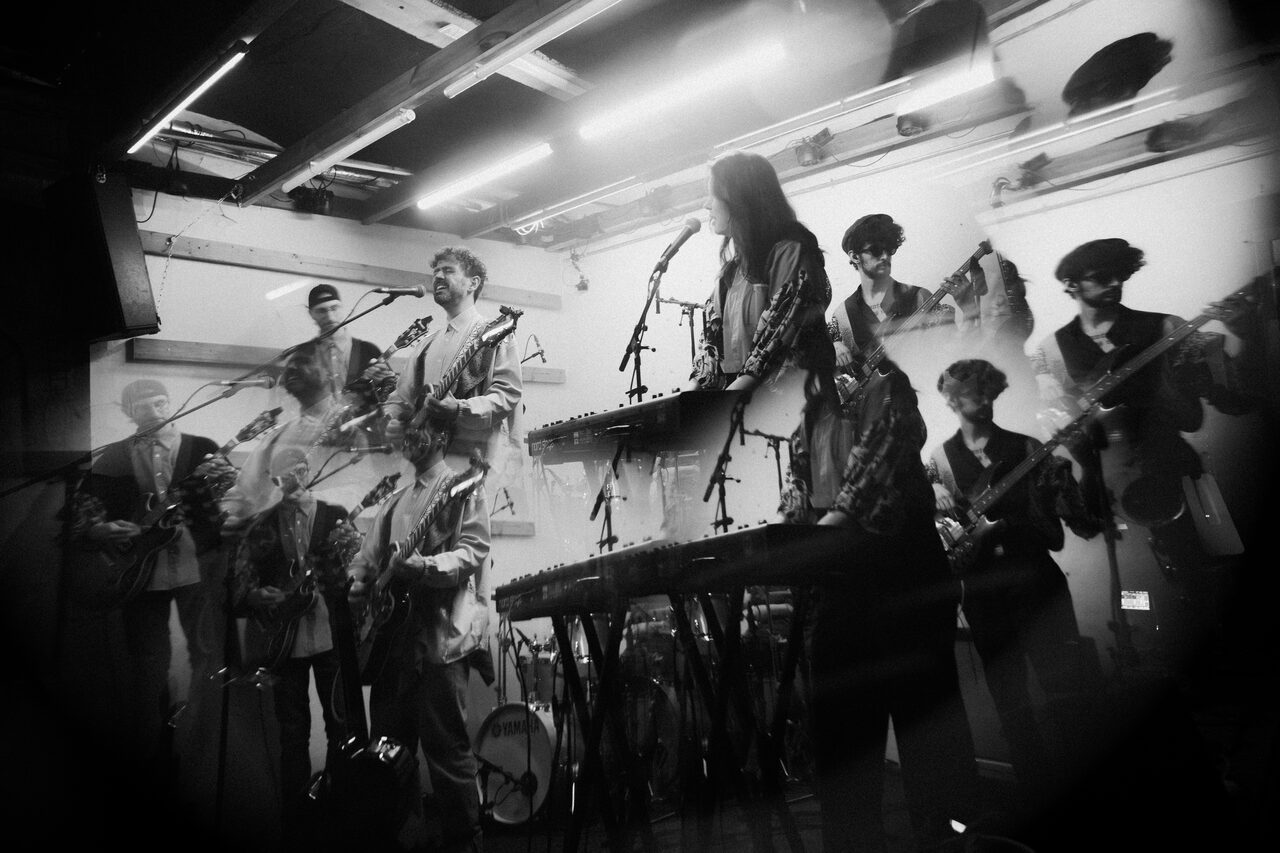 The band playing on a small stage with keyboards and guitars, in a black-and-white double exposure.